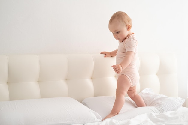 Bebê fofo em um carrinho de macacão segurando a cabeceira da cama e aprendendo a andar na cama branca