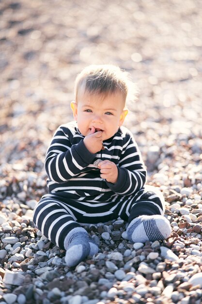 Bebê fofo em um macacão listrado sentado em uma praia de seixos segurando uma pedra contra a boca
