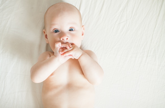 Bebê fofo encontra-se em um lençol branco e brinca alegremente, mordisca as mãos