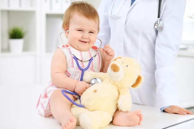 Foto bebê fofo feliz no exame de saúde no consultório do médico a menina da criança está sentada e mantendo o estetoscópio e o ursinho de pelúcia