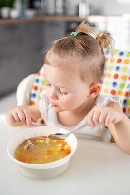 Bebê fofo menina sentada na cadeira alta e comendo sua sopa de almoço na cozinha de casa