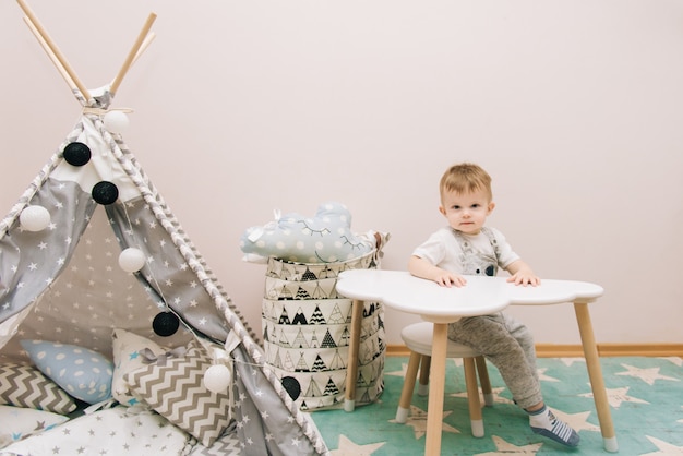 Bebê fofo sentado à mesa no quarto das crianças em tons de brancos, cinza e azuis. perto da tenda e um saco de brinquedos