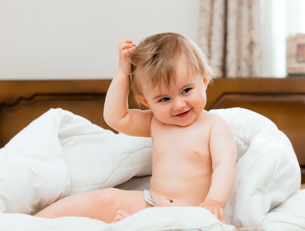 Foto bebê fofo sentado em uma cama, enrolado em um cobertor branco. bebê tem 11 meses.