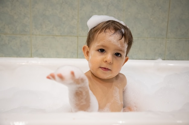 Bebé fofo tomando banho olha para a câmera e segura a espuma de sabão perto do foco suave