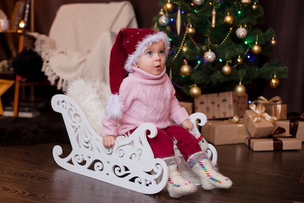 Bebé con un gorro de Navidad con un suéter y calcetines de lana en un trineo en un interior de Año Nuevo Emociones