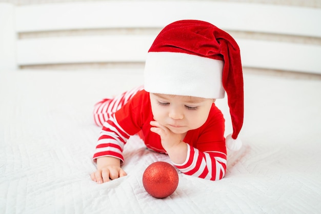 Bebé con gorro de Papá Noel en bola de Navidad en la cama vacaciones de año nuevo santa bebé gracioso