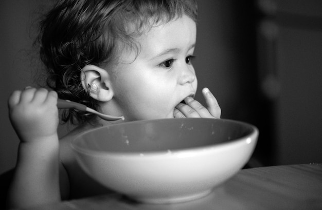 Bebé gracioso comiendo comida él mismo con una cuchara en la cocina