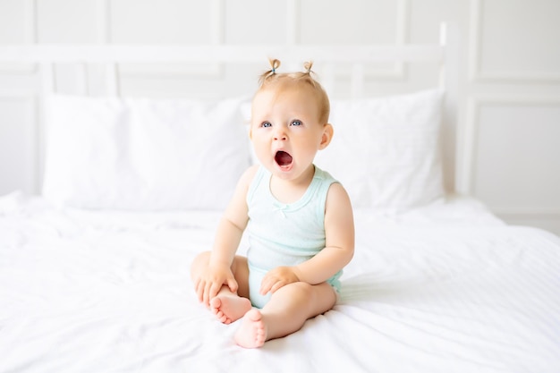 Un bebé gracioso parado en una cuna en una guardería blanca con una ventana Una niña con un traje de algodón El bebé bosteza quiere dormir preguntas sobre el sueño de los niños