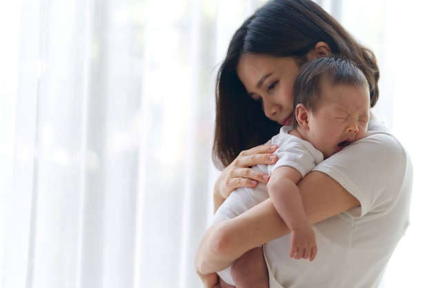 El bebé infantil asiático lindo en las madres da la situación en el cuarto cerca de la ventana.