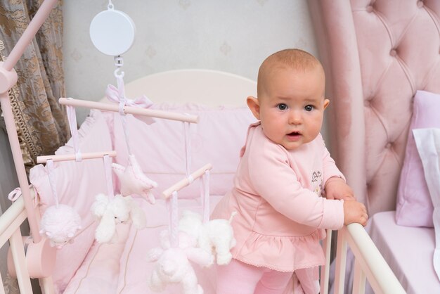 Un bebé infantil feliz y curioso en una cuna rosa y una escena de dormitorio con un móvil colgante.