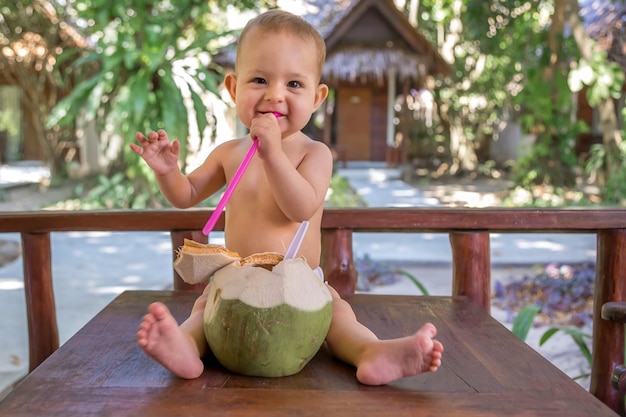 Bebê infantil feliz em férias tropicais Come e bebe coco verde jovem Senta-se no chão