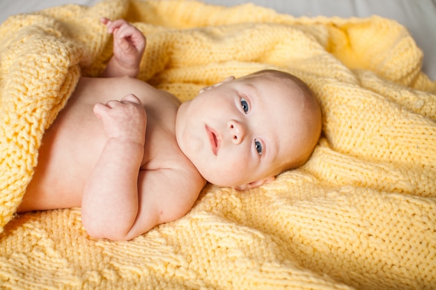 Bebé infantil lindo que mira la cámara en una tela escocesa amarilla hecha punto en una cama blanca.