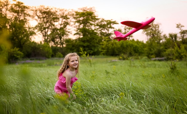 El bebé lanza un avión rosa en el fondo del bosque y la hierba alta. Jugando con un avión rosa con un traje rosa