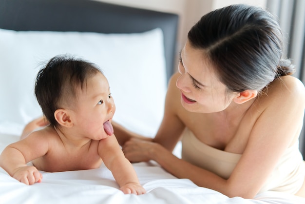 Bebé lindo asiático que se arrastra y que juega con su madre hermosa en la cama.