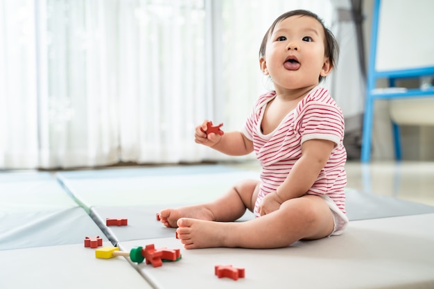 Bebé lindo asiático que sienta y que juega un pequeño juguete en la estera suave en casa.