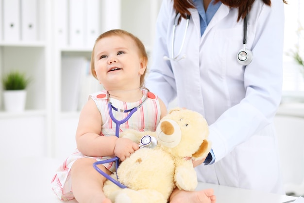 Bebé lindo feliz en el examen de salud en el consultorio del médico Niña pequeña está sentada y manteniendo el estetoscopio y el oso de peluche