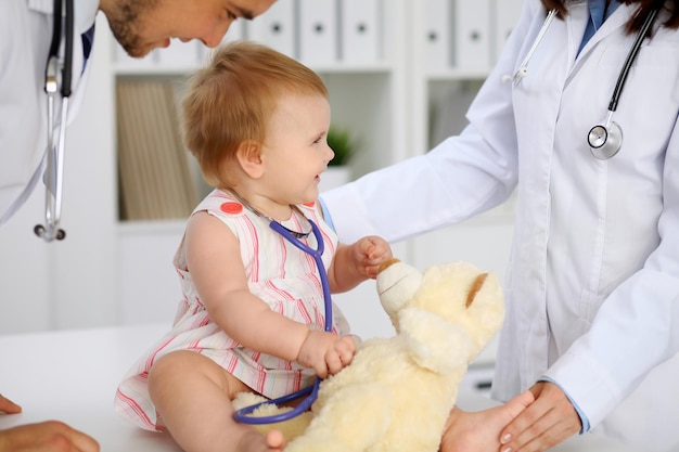 Bebé lindo feliz en el examen de salud en el consultorio del médico Niña pequeña está sentada y manteniendo el estetoscopio y el oso de peluche