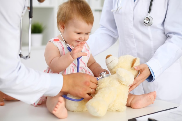 Bebé lindo feliz en el examen de salud en el consultorio del médico Niña pequeña está sentada y manteniendo el estetoscopio y el oso de peluche