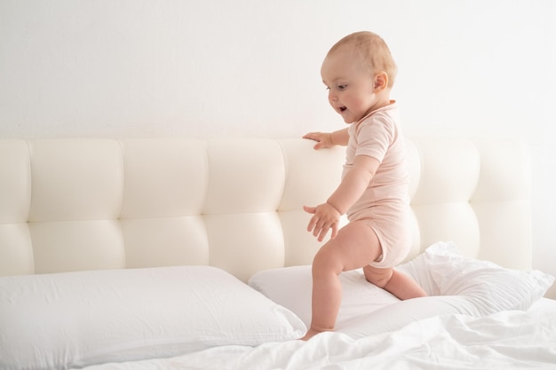 Bebé lindo en soporte del mono que sostiene el marco de la cama que aprende a caminar en la ropa de cama blanca en la cama