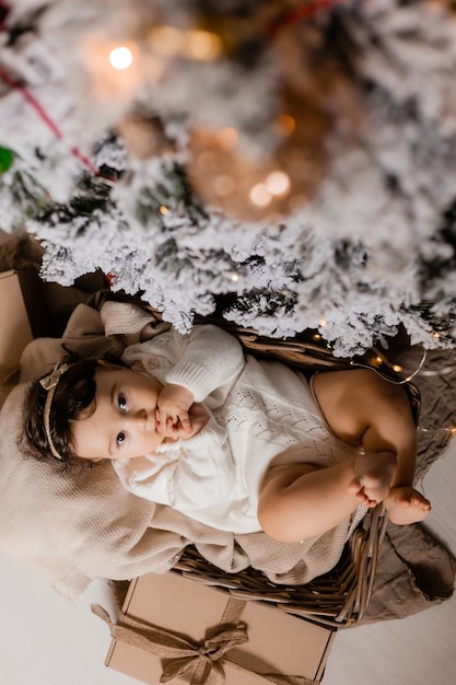 un bebé lindo en traje blanco yace en una cuna junto al árbol de Navidad