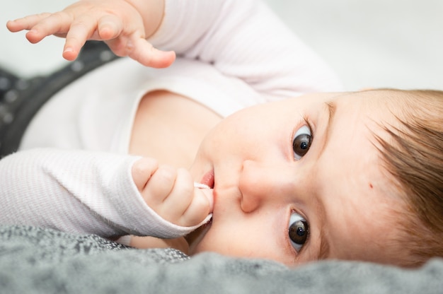 Bebê louro adorável que encontra-se no seu para trás na cama branca que olha diretamente na câmera.