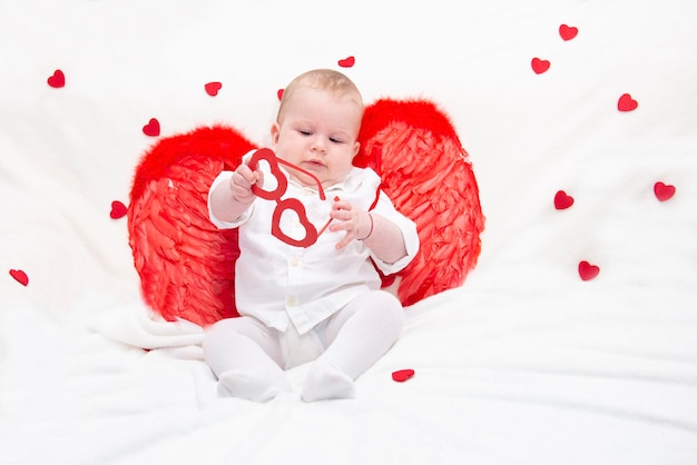 Un bebé con mejillas hinchadas en ropa blanca con alas rojas de cupido juega con gafas con corazones aislados