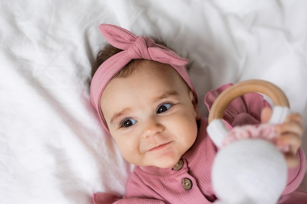 El bebé en un mono rosa con un cascabel de madera ecológico está tendido en la cama