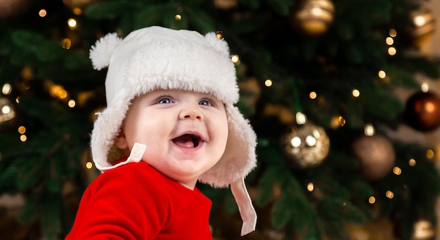 El bebé de Navidad está sonriendo. La niña linda con un vestido rojo y un sombrero blanco expresa emociones. Concepto de Navidad con niño