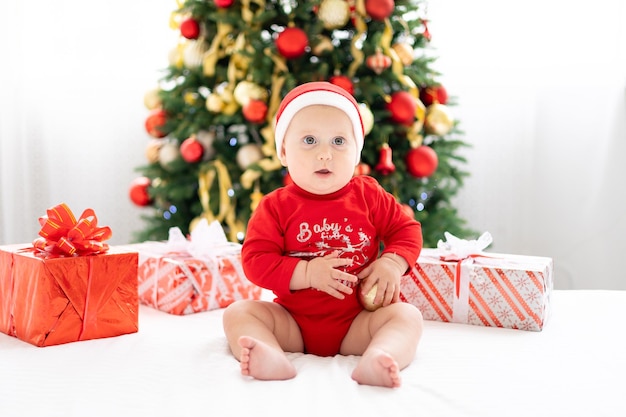 Bebé niño feliz en traje rojo de santa celebrando el año nuevo en casa con árbol de navidad y regalos, concepto de vacaciones, feliz navidad y año nuevo