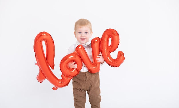 Bebé niño en traje tiene la inscripción amor de globos en una pared blanca.