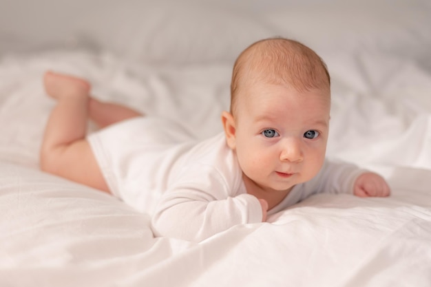 Bebé con ojos azules en un traje blanco yace boca abajo sobre ropa de cama blanca. estilo de vida. espacio para texto. foto de alta calidad