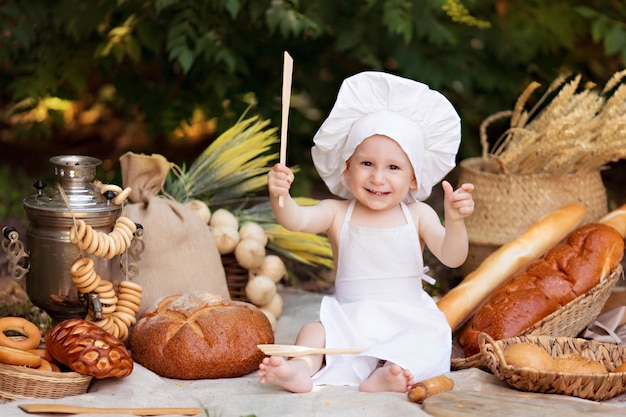 Bebé panadero cocina en la naturaleza. niño en un picnic come pan y rosquillas en un delantal blanco y sombrero