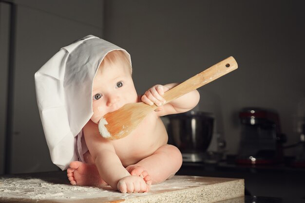 Bebé panadero en sombrero de cocinero con una cuchara en la boca en la cocina