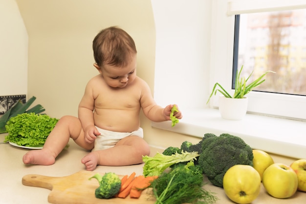 El bebé en el pañal está sentado en la mesa de la cocina. Un niño juega y se divierte con frutas y verduras orgánicas frescas.