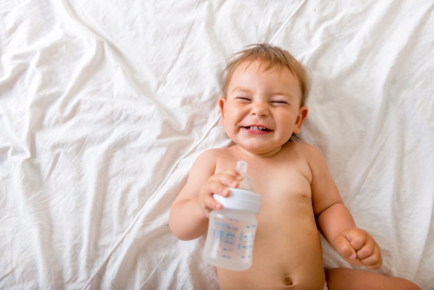 Foto bebé pequeño acostado en la cama blanca, sonríe y bebe agua de una botella de plástico.