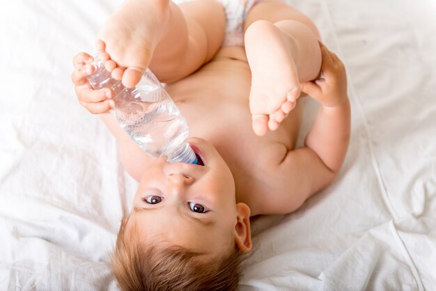Bebé pequeño acostado en la cama blanca, sonríe y bebe agua de una botella de plástico.