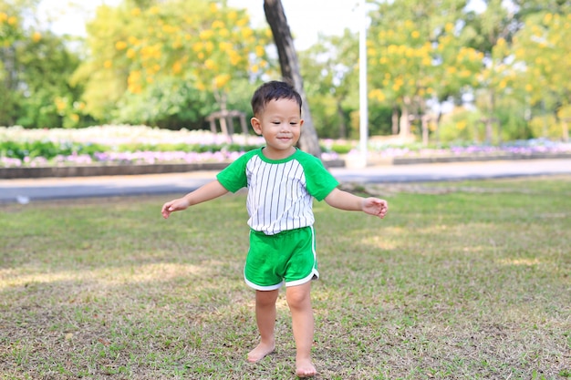 Bebê pequeno asiático feliz que anda com os pés descalços na grama no jardim do verão.