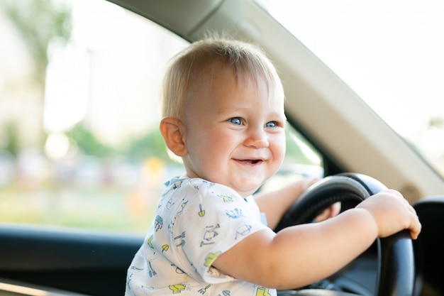 Bebé pequeno bonito que conduz o carro grande, guardando o volante, sorrindo e olhando para a frente com interesse.