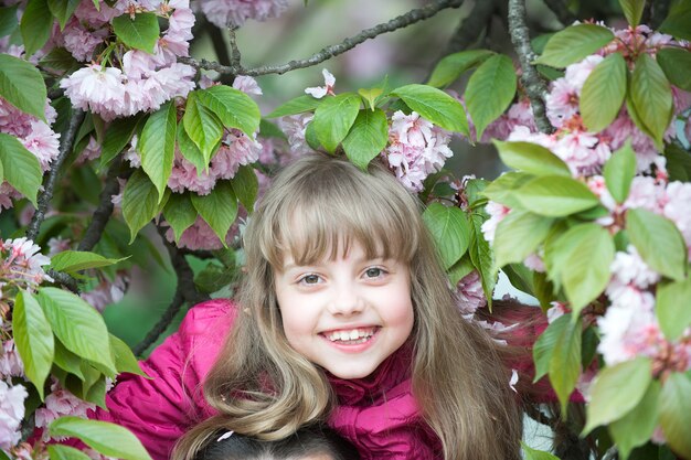 Bebé pequeno ou criança fofa com adorável rosto sorridente e cabelo loiro em uma camisa rosa entre flor de sakura primavera, flor de cerejeira com folhas verdes ao ar livre em fundo natural