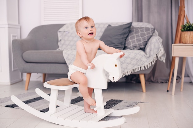 Bebê pequeno sorridente, jogando em um cavalo de madeira do brinquedo na sala aconchegante.