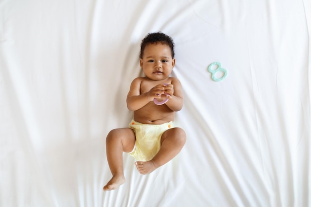 Bebê preto usando fralda reutilizável brincando com brinquedo enquanto estava deitado na cama