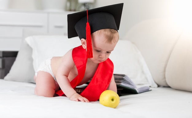 Bebé prodigio en gorro de graduación y cinta roja para alcanzar la manzana