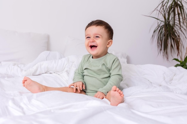 El bebé que se ríe con un traje de algodón verde y los pies descalzos está sentado en una cama de alta calidad