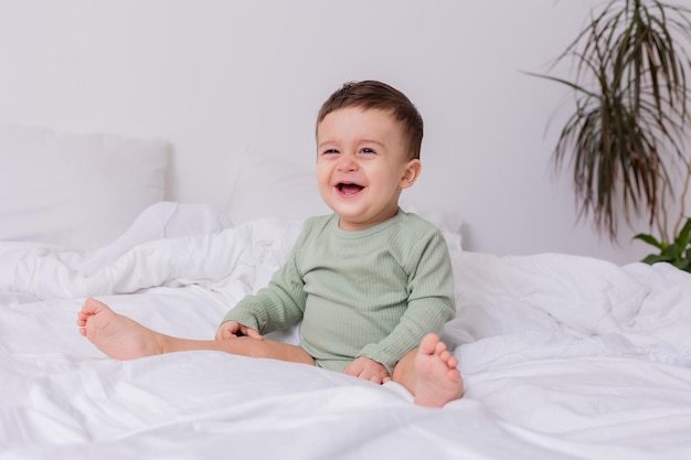 El bebé que se ríe con un traje de algodón verde y los pies descalzos está sentado en una cama de alta calidad