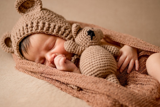 Bebê recém-nascido, 10 dias de idade, dormindo com uma roupa aconchegante de urso e com uma roupa marrom claro. Ele está segurando um pequeno ursinho de pelúcia. Começo da vida e o conceito de infância feliz.