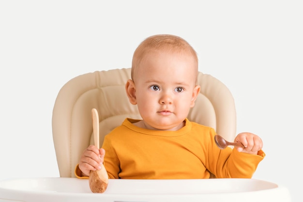 Bebê recém-nascido de 12 a 17 meses segura colheres de madeira enquanto está sentado em uma cadeira de alimentação