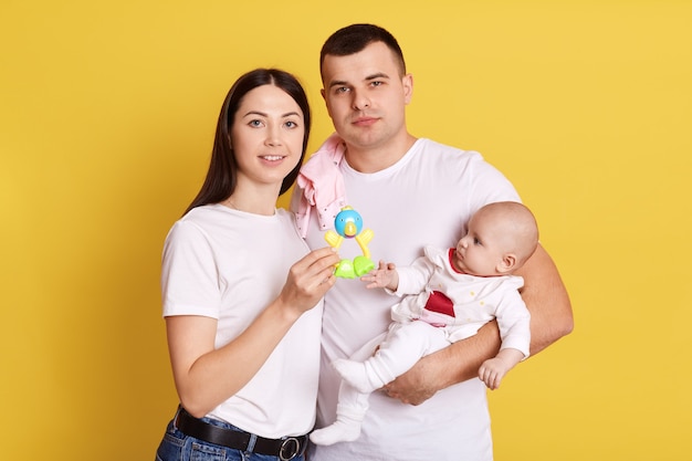 Bebê recém-nascido e pais vestindo fundo amarelo, família vestindo camisetas brancas, olhando para a câmera, mãe mostrando o pufe para criança, família feliz, passando algum tempo juntos.