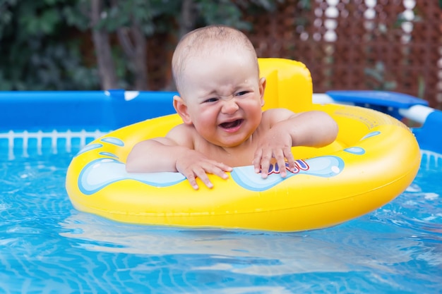 Un bebé recién nacido de 10 a 12 meses que llora nada en un anillo de natación amarillo. Piscina de marco en el patio trasero de la casa