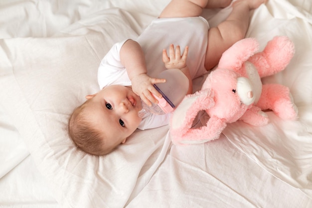 Un bebé recién nacido alegre con un traje blanco yace en una cama blanca con un conejo de peluche rosa. productos para niños, juguete. concepto de una infancia feliz y maternidad. cuidado de los niños. espacio para texto.