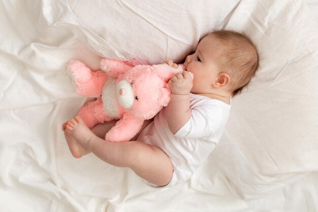 Un bebé recién nacido alegre con un traje blanco yace en una cama blanca con un conejo de peluche rosa. productos para niños, juguete. concepto de una infancia feliz y maternidad. cuidado de los niños. espacio para texto.